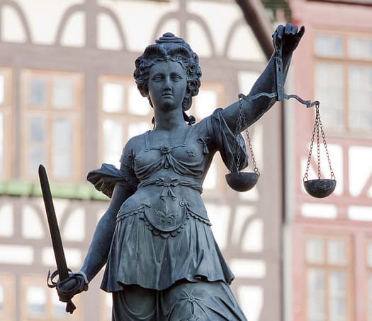 Teisingumo deivė vaizduojama akla, kairėje laikanti svarstykles, dešinėje – kalaviją (teisingumui įgyvendinti)